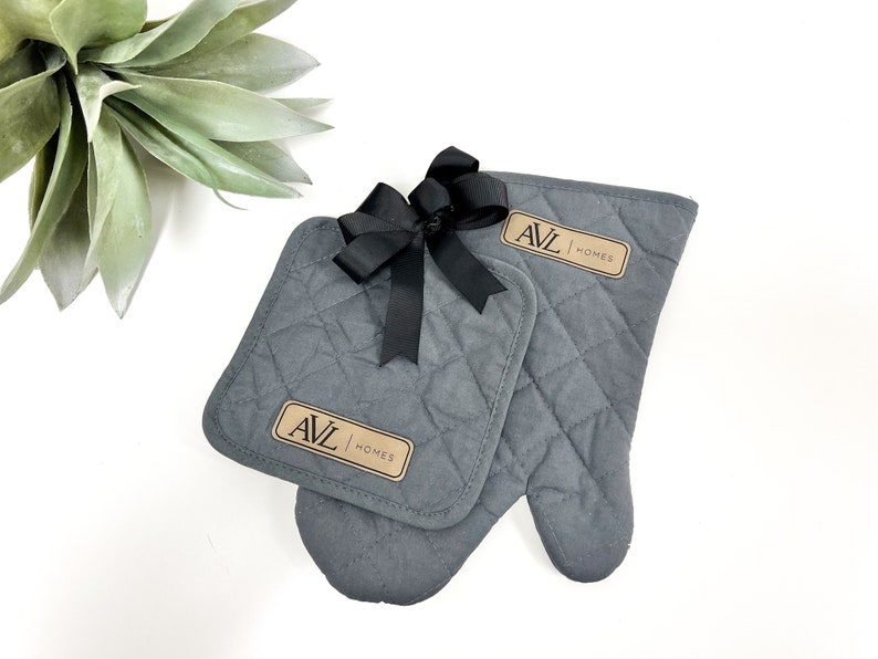 May include: A set of two gray quilted oven mitts with a brown label that says "AVL HOMES". The mitts are tied together with a black ribbon.