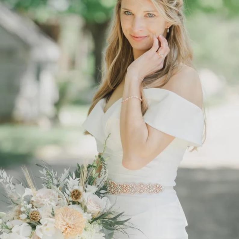 May include: A white off-the-shoulder dress featuring a wide, ornate rose gold metallic belt with intricate filigree detailing. The garment is paired with a delicate rose gold bracelet and a large bouquet of white and peach-toned flowers.
