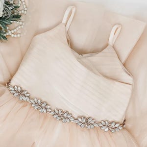 May include: A champagne-coloured dress featuring a sleeveless bodice with horizontal pleats and thin shoulder straps. The waistline is accented with a decorative band of clear, marquise-cut crystals arranged in a floral pattern over a tulle skirt.