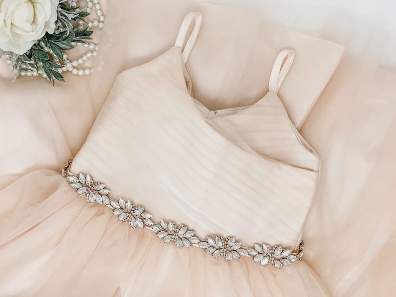 May include: A champagne-coloured dress featuring a sleeveless bodice with horizontal pleats and thin shoulder straps. The waistline is accented with a decorative band of clear, marquise-cut crystals arranged in a floral pattern over a tulle skirt.