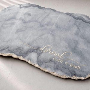 May include: An oval-shaped gray and white marbled serving board with a textured, metallic gold-colored edge. The surface features the gold-colored script text "dornak" above the names "CODY" and "JAMIE" separated by a small heart symbol.