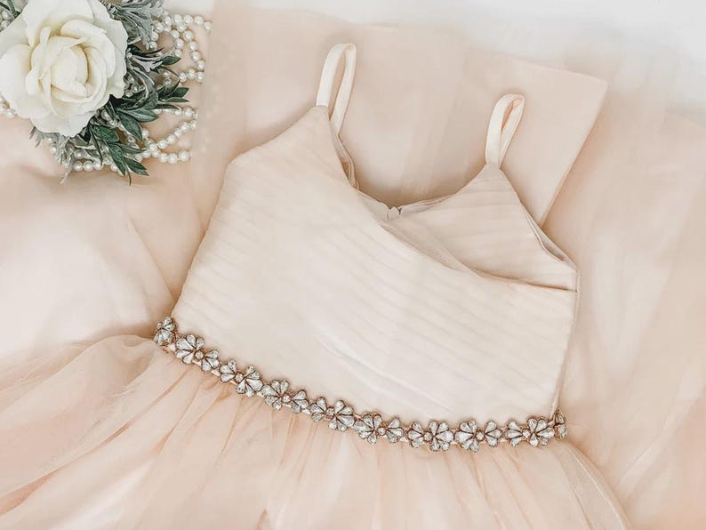 May include: A pale blush pink dress featuring a sleeveless bodice with thin straps and a horizontal pleated texture. The waistline is accented with a decorative band of clear, flower-shaped crystals. The skirt is made of layered, sheer tulle fabric.