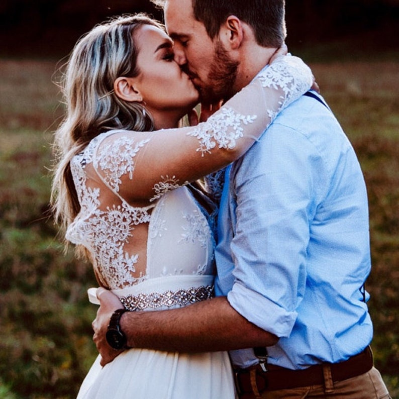May include: A white wedding dress with long lace sleeves and a beaded waist belt paired with a light blue button-down shirt, brown trousers, and dark braces. The dress features intricate floral lace detailing on the bodice and sleeves.