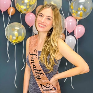 Puede incluir: Una mujer con un vestido gris brillante con una banda de color rosa dorado que dice "Finalmente 21". Est&aacute; de pie frente a una pared decorada con globos rosas y transparentes.