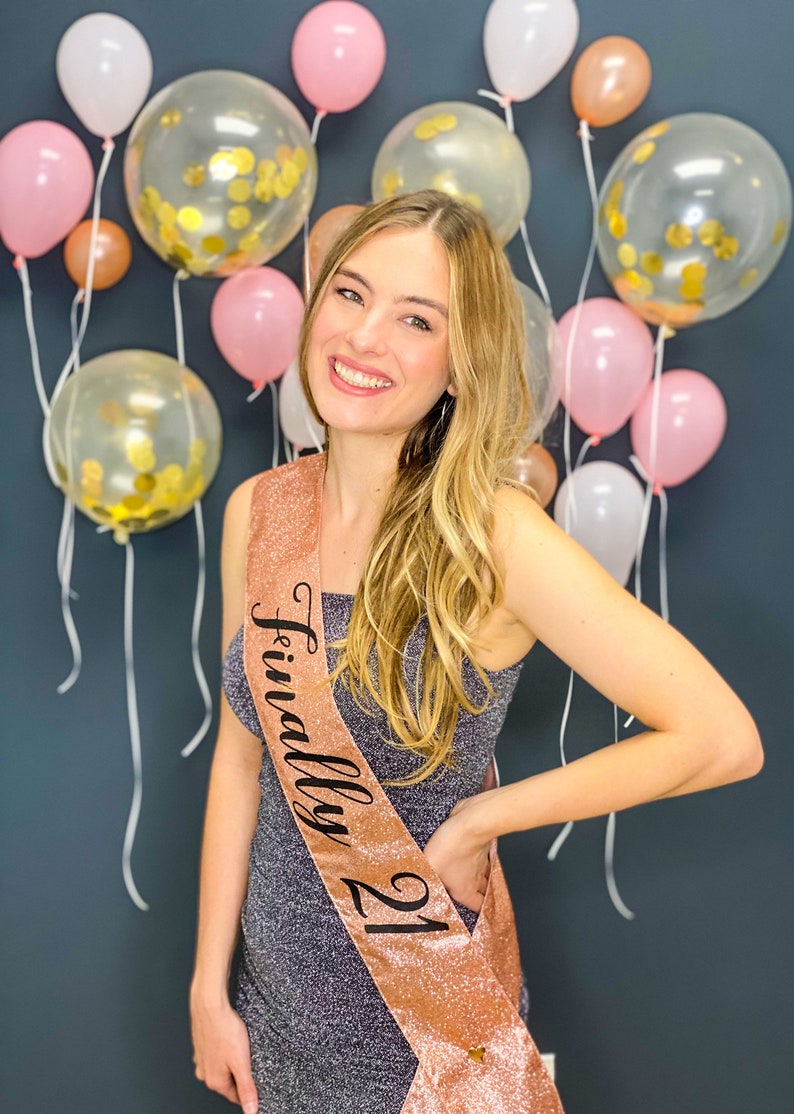 Puede incluir: Una mujer con un vestido gris brillante con una banda de color rosa dorado que dice "Finalmente 21". Est&aacute; de pie frente a una pared decorada con globos rosas y transparentes.