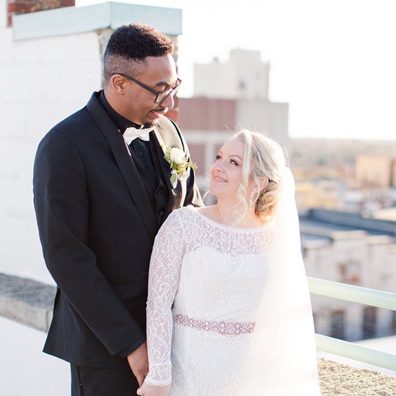 May include: A black suit jacket with a white bow tie and a white floral boutonniere paired with a white long-sleeved lace wedding dress featuring a decorative rose gold waist belt and a sheer veil, captured in a bright outdoor setting.