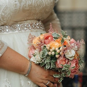 May include: A bouquet of peach, pink, and white roses mixed with white berries and feathery pink astilbe, held against a cream-coloured dress featuring an intricate white embroidered pattern and a wide, sparkling rhinestone belt.