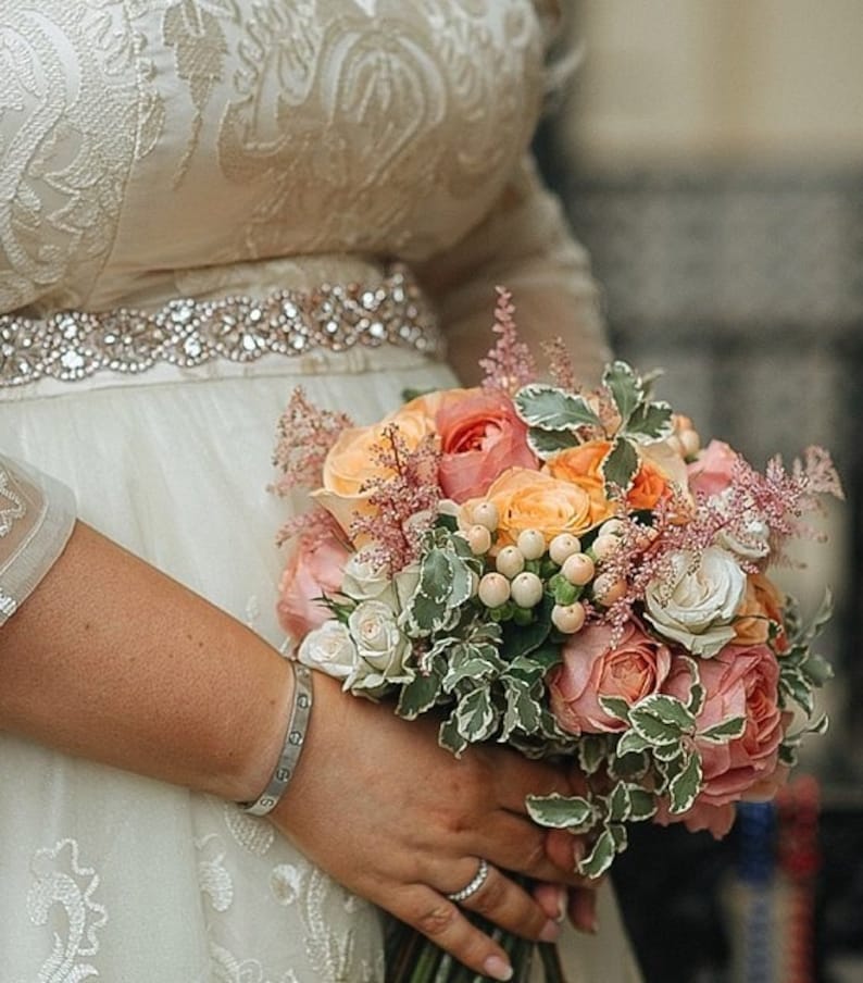 May include: A bouquet of peach, pink, and white roses mixed with white berries and feathery pink astilbe, held against a cream-coloured dress featuring an intricate white embroidered pattern and a wide, sparkling rhinestone belt.