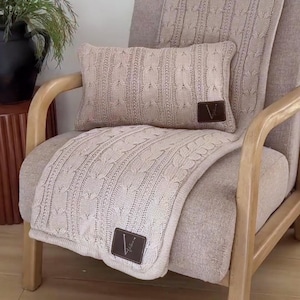 May include: A beige cable-knit rectangular pillow and a matching cable-knit throw blanket draped over a chair. Both items feature a dark brown rectangular leather patch with the letter V embossed in white, adding a distinct detail to the set.
