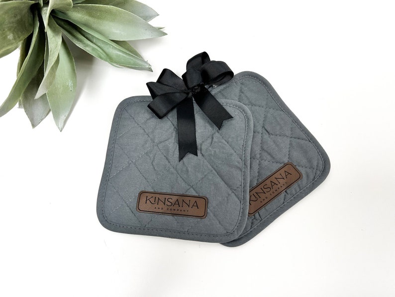 May include: Two gray quilted pot holders with a black bow and a brown leather label that says "Kinsana and Company".