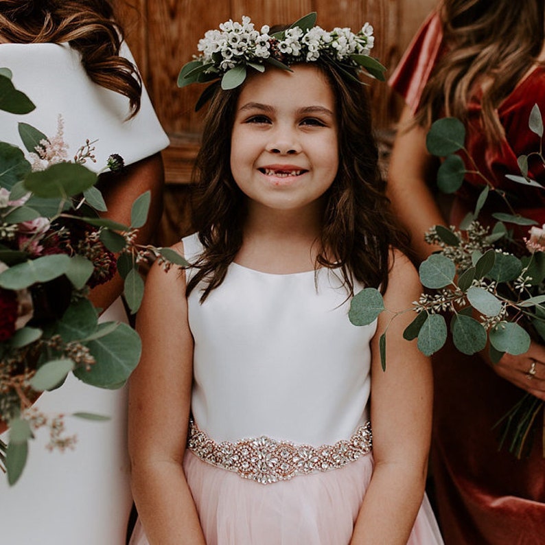 May include: A sleeveless dress with a white bodice and a pale pink tulle skirt, featuring a wide, rose gold-toned belt embellished with clear crystals. The dress is worn with a floral crown made of small white blossoms and green leaves.