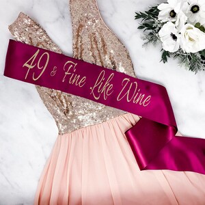 Puede incluir: Una banda de sat&eacute;n burdeos con letras doradas brillantes que dicen "49 & Fine Like Wine". La banda est&aacute; envuelta alrededor de un vestido de lentejuelas rosa y dorado.