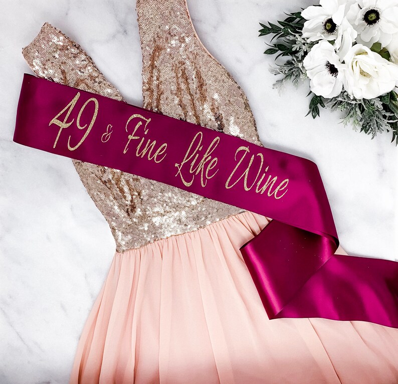 Puede incluir: Una banda de sat&eacute;n burdeos con letras doradas brillantes que dicen "49 & Fine Like Wine". La banda est&aacute; envuelta alrededor de un vestido de lentejuelas rosa y dorado.