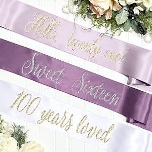 Puede incluir: Tres fajas de sat&eacute;n con letras doradas de purpurina. La faja superior es lavanda y dice "Hello, twenty one!" La faja del medio es morada y dice "Sweet Sixteen." La faja inferior es blanca y dice "100 years loved."