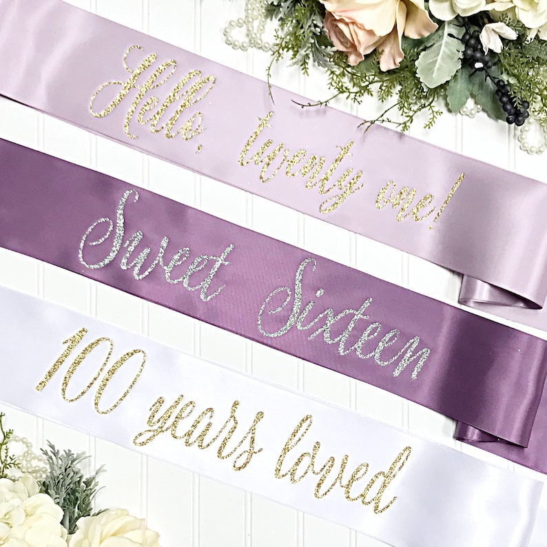 Puede incluir: Tres fajas de sat&eacute;n con letras doradas de purpurina. La faja superior es lavanda y dice "Hello, twenty one!" La faja del medio es morada y dice "Sweet Sixteen." La faja inferior es blanca y dice "100 years loved."