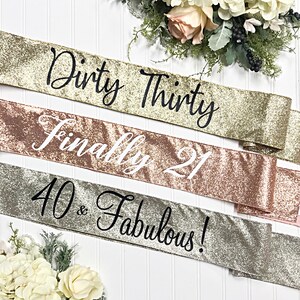 Puede incluir: Tres fajas doradas con purpurina y texto negro. La faja superior dice "Dirty Thirty", la faja del medio dice "Finally 21" y la faja inferior dice "40 & Fabulous!"