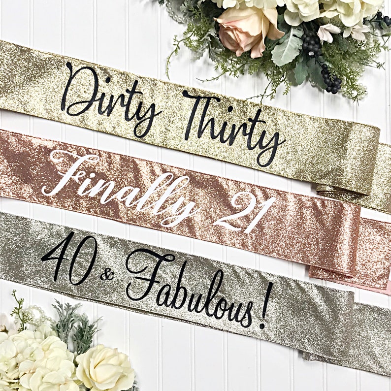 Puede incluir: Tres fajas doradas con purpurina y texto negro. La faja superior dice "Dirty Thirty", la faja del medio dice "Finally 21" y la faja inferior dice "40 & Fabulous!"