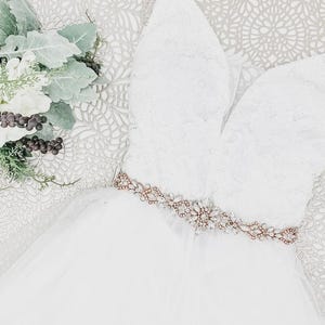 May include: A white sleeveless dress featuring a lace bodice with a deep V-neckline and a tulle skirt. A rose gold belt adorned with clear crystals and small white pearls is cinched at the waist. A bouquet of white flowers and green leaves is nearby.