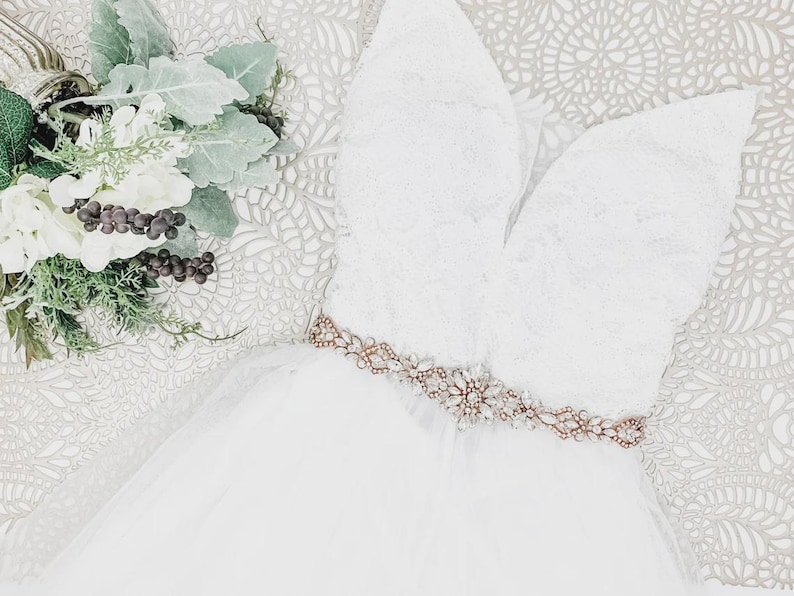 May include: A white sleeveless dress featuring a lace bodice with a deep V-neckline and a tulle skirt. A rose gold belt adorned with clear crystals and small white pearls is cinched at the waist. A bouquet of white flowers and green leaves is nearby.
