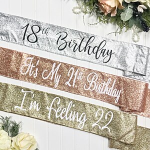 Puede incluir: Tres fajas brillantes para una celebraci&oacute;n de cumplea&ntilde;os. Las fajas son plateadas, oro rosa y doradas. El texto en las fajas dice "18th Birthday", "It's My 21st Birthday", y "I'm Feeling 22".