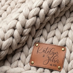 May include: A chunky knit blanket in a light beige color featuring a rectangular tan leather patch attached with four brass-colored rivets. The patch is engraved with the text Katelyn & John and est. 2026 in a black script and sans-serif font.