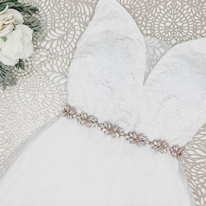 May include: A white sleeveless dress featuring a lace bodice with a deep V-neckline and a tulle skirt. The waist is accented with a metallic belt adorned with clear crystals and floral-shaped embellishments. A cluster of white flowers sits nearby.