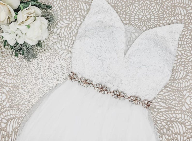 May include: A white sleeveless dress featuring a lace bodice with a deep V-neckline and a tulle skirt. The waist is accented with a metallic belt adorned with clear crystals and floral-shaped embellishments. A cluster of white flowers sits nearby.