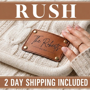 May include: A rectangular tan leather patch with rounded corners and four brass-colored rivets attached to a cream-colored cable knit fabric. The patch features the black text "The Roberts" and "EST. 2026" centered on its surface.