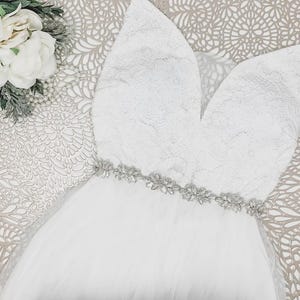 May include: A white sleeveless dress featuring a deep V-neckline with floral lace detailing on the bodice. The waist is accented by a horizontal band of clear, sparkling crystals and silver-toned embellishments, transitioning into a full tulle skirt.