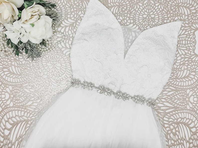 May include: A white sleeveless dress featuring a deep V-neckline with floral lace detailing on the bodice. The waist is accented by a horizontal band of clear, sparkling crystals and silver-toned embellishments, transitioning into a full tulle skirt.