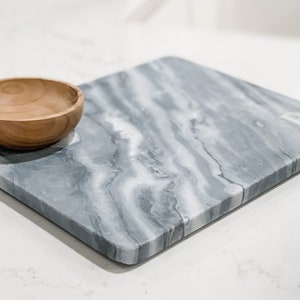 May include: A gray marble serving board with a white swirl pattern. The board has a beveled edge and a small wooden bowl on top.