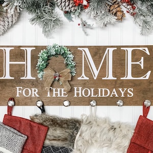 May include: Wooden sign with the words "HOME FOR THE HOLIDAYS" in white. A small wreath and bow are attached to the sign. Below the sign are metal hooks with Christmas stockings hanging from them.