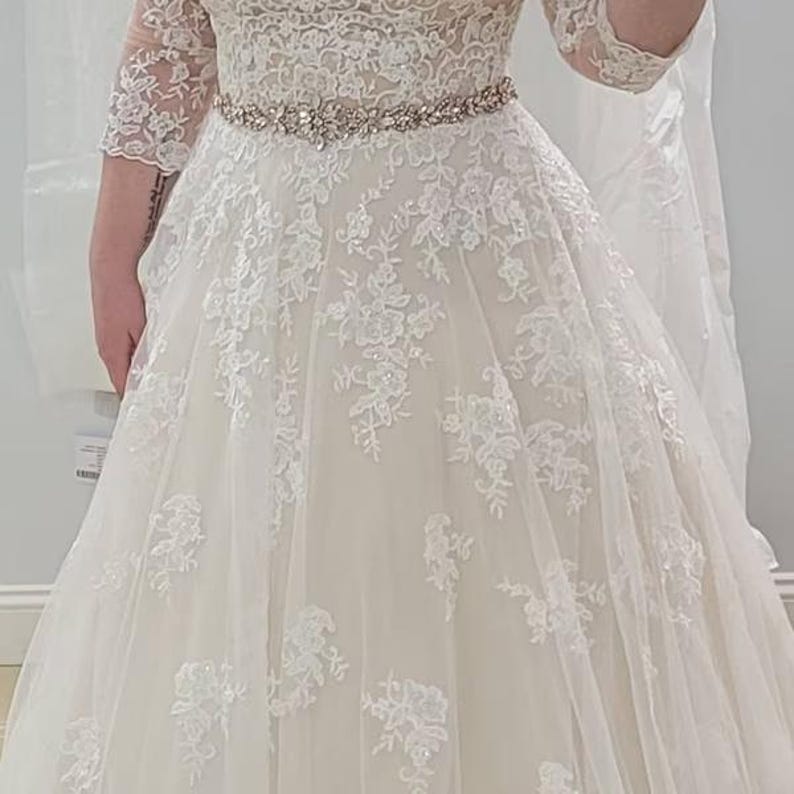 May include: A floor-length ivory gown featuring three-quarter length lace sleeves and a bodice with intricate floral lace embroidery. The waist is accented by a thin, sparkling rhinestone belt, and the skirt consists of layered tulle fabric.