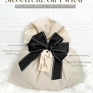 a beige gift bag with a black bow and a tag that says "Home Sweet Home". The bag is described as a "Signature Gift Wrap" and is made of a reusable canvas bag.