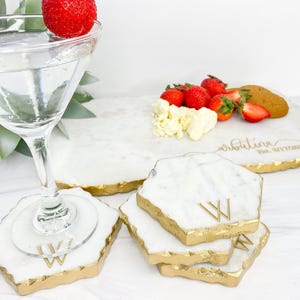 May include: A set of white marble coasters and a rectangular serving board, each featuring irregular edges finished in gold. The coasters are engraved with the letter W, while the board displays the script text arbutina and Est. 8/17/2017.