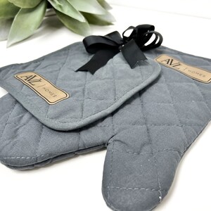 May include: A pair of gray quilted oven mitts with a black ribbon bow. The mitts have a brown leather patch with the text "AVL HOMES" on them.