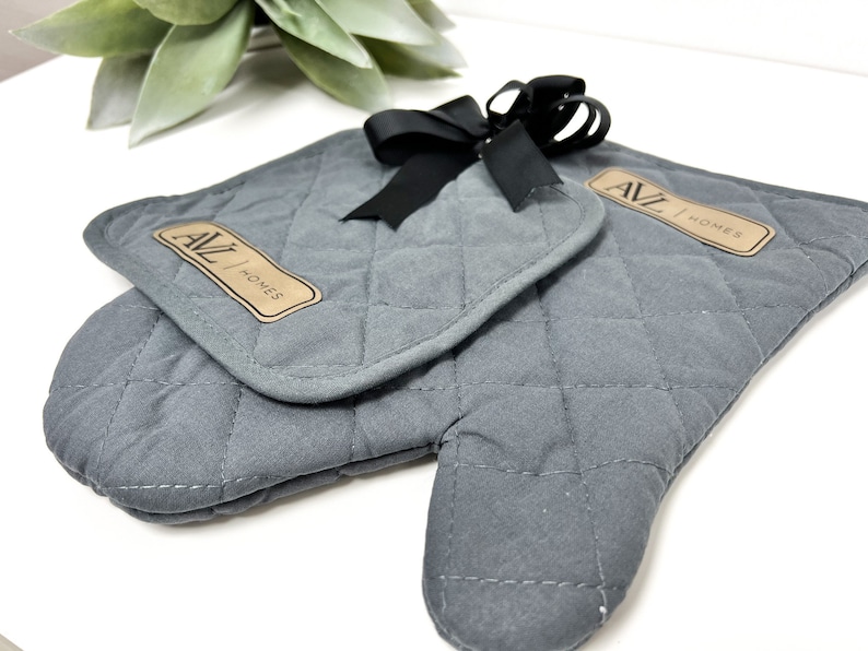 May include: A pair of gray quilted oven mitts with a black ribbon bow. The mitts have a brown leather patch with the text "AVL HOMES" on them.