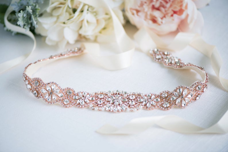 May include: A rose gold belt featuring a central floral pattern embellished with clear crystals and small rhinestones. The band is constructed from a metallic rose gold base and includes long, smooth cream-coloured satin ribbons attached to each end.