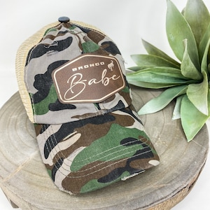 Puede incluir: Una gorra de camionero de camuflaje con un parche de cuero marrón que dice "Bronco Babe" en cursiva.