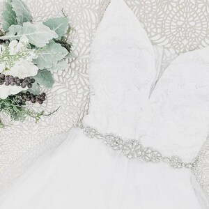 May include: A white sleeveless dress featuring a deep V-neckline, a bodice with intricate lace detailing, and a waistline embellished with a silver-toned crystal belt. The skirt is made of layered tulle fabric, accompanied by a bouquet of white flowers.