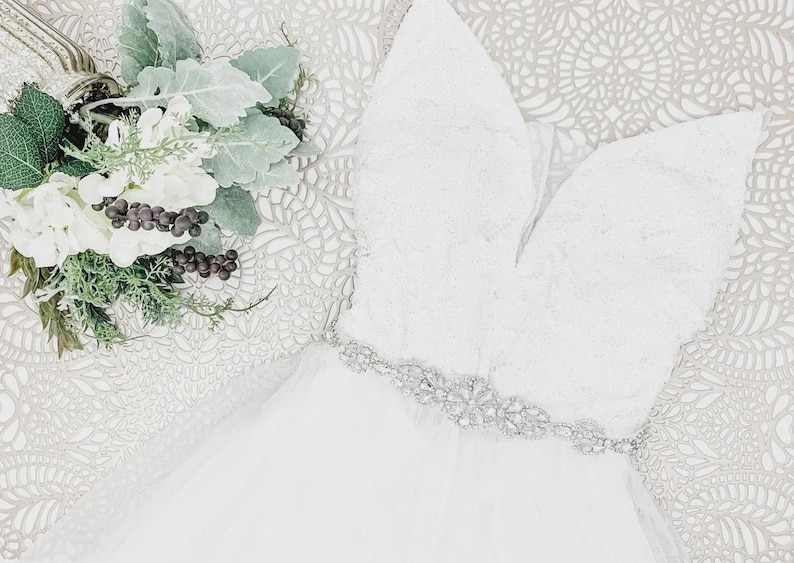 May include: A white sleeveless dress featuring a deep V-neckline, a bodice with intricate lace detailing, and a waistline embellished with a silver-toned crystal belt. The skirt is made of layered tulle fabric, accompanied by a bouquet of white flowers.