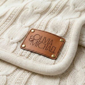 Op de afbeelding: Een gebroken witte kabelgebreide deken met een rechthoekige lichtbruine leren patch die is bevestigd met vier messingkleurige metalen klinknagels. De leren patch is voorzien van de tekst OLIVIA MICHAEL in een modern schreefloos lettertype.