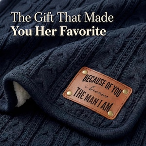 May include: A navy blue cable-knit blanket with a soft, white sherpa lining. A brown leather patch is attached, reading "BECAUSE OF YOU i became THE MAN I AM". The text "The Gift That Made You Her Favorite" is at the top.