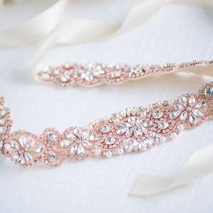 May include: A rose gold belt featuring a repeating floral pattern encrusted with clear faceted crystals and small white pearls. The decorative section is attached to long, smooth, cream-coloured satin ribbons on both ends for tying.