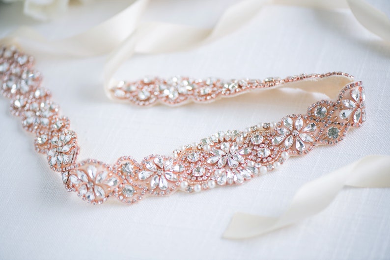 May include: A rose gold belt featuring a repeating floral pattern encrusted with clear faceted crystals and small white pearls. The decorative section is attached to long, smooth, cream-coloured satin ribbons on both ends for tying.