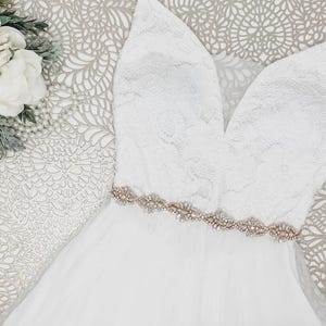 May include: A white sleeveless dress featuring a lace bodice with a deep V-neckline and a flowing skirt. A metallic rose gold belt adorned with clear crystals and beads sits at the waist. A cluster of white artificial flowers is visible nearby.