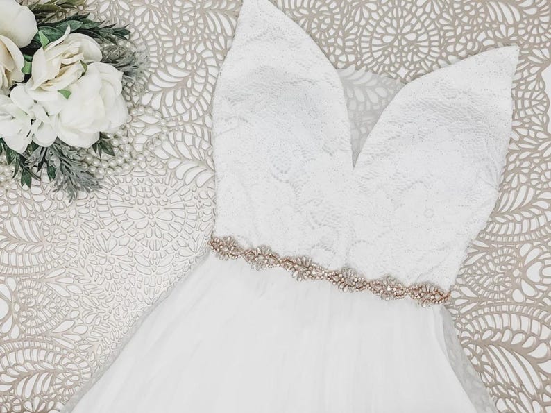 May include: A white sleeveless dress featuring a lace bodice with a deep V-neckline and a flowing skirt. A metallic rose gold belt adorned with clear crystals and beads sits at the waist. A cluster of white artificial flowers is visible nearby.