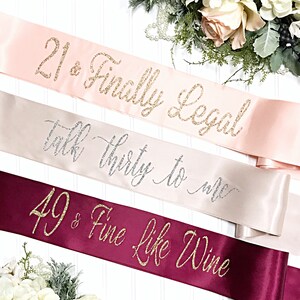 Puede incluir: Tres fajas de sat&eacute;n con letras doradas de purpurina. La faja superior es rosa con el texto "21 & Finalmente Legal". La faja del medio es beige con el texto "Habla Treinta Conmigo". La faja inferior es burdeos con el texto "49 & Fino Como El Vino".