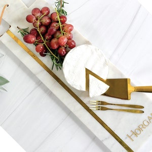May include: A white marble serving board with a gold trim and the name "HORGAN" engraved on it. The board is decorated with a wedge of brie cheese, a bunch of red grapes, and rosemary sprigs. A gold cheese knife and two gold forks are also included.