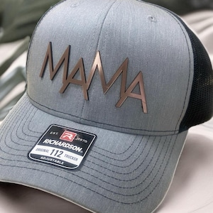 Może przedstawiać: Szara i czarna czapka trucker z logo "MAMA" w kolorze różowego złota. Czapka jest regulowana i ma metkę Richardson 112.